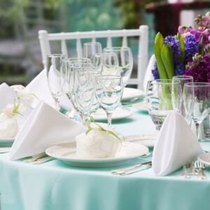 A beautifully set table with a light blue tablecloth features white napkins folded neatly, multiple wine glasses, and place settings. The centerpiece includes purple flowers and green tulips, creating an elegant and inviting atmosphere, complemented by thoughtfully chosen event furniture.
