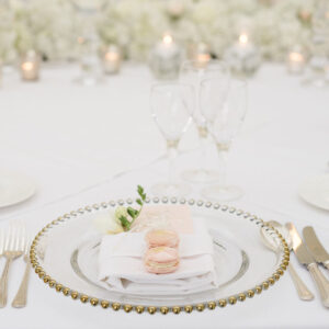 A place setting at a formal event features a glass plate with gold beading, holding a white napkin and two macarons. Surrounding the plate are silver cutlery, elegant event furniture with white flowers as centerpieces, and several empty wine glasses.