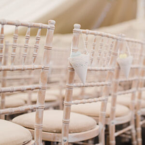 Wooden chairs with beige cushions are lined up in rows. Each chair has a small, white, lace-trimmed cone hanging from the backrest, likely for holding petals or confetti. The setting appears to showcase elegant event furniture, possibly for a wedding.